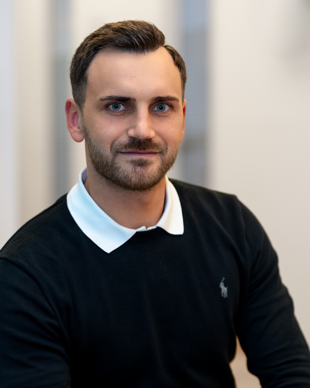 Person steht in einem hellen Innenraum und trägt einen schwarzen Pullover mit weißem Hemdkragen. Ein Arm ist entspannt auf einer Oberfläche abgestützt.