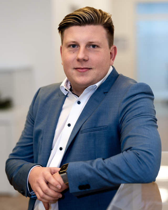 Person in blauem Sakko und weißem Hemd lehnt sich leicht über eine Glasfläche in einem modernen Büro.